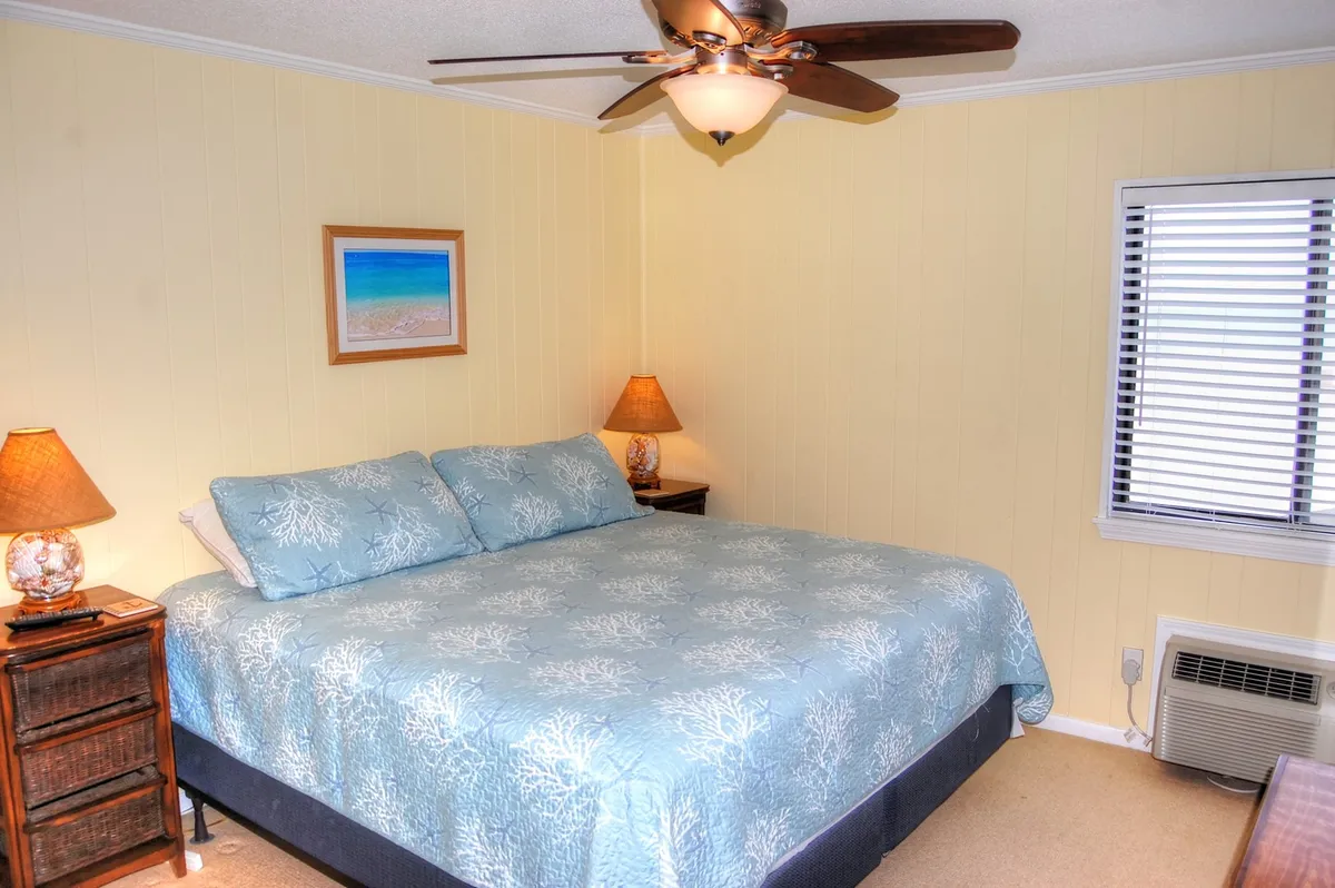 A serene bedroom featuring a queen-sized bed with light blue bedding, flanked by nightstands and lamps. Above the bed hangs a framed beach painting, and natural light streams in, creating a calm, inviting ambiance. A ceiling fan adds an element of comfort.