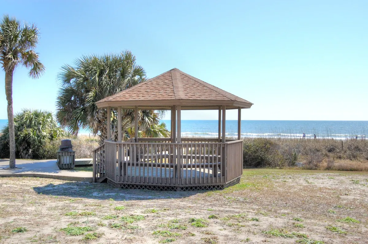 A tranquil beachfront scene featuring a wooden gazebo overlooking the ocean, framed by swaying palm trees and soft sand. The setting is ideal for couples to enjoy peaceful moments, whether it's sipping coffee at sunrise or sharing a sunset together in privacy.