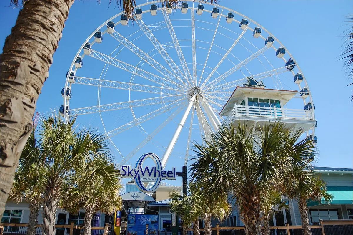 This image features the iconic SkyWheel and its entrance surrounded by palm trees, capturing visitors in the joyful act of entering or photographing the landmark. The SkyWheel dominates the scene, illustrating a celebrated Myrtle Beach attraction.
