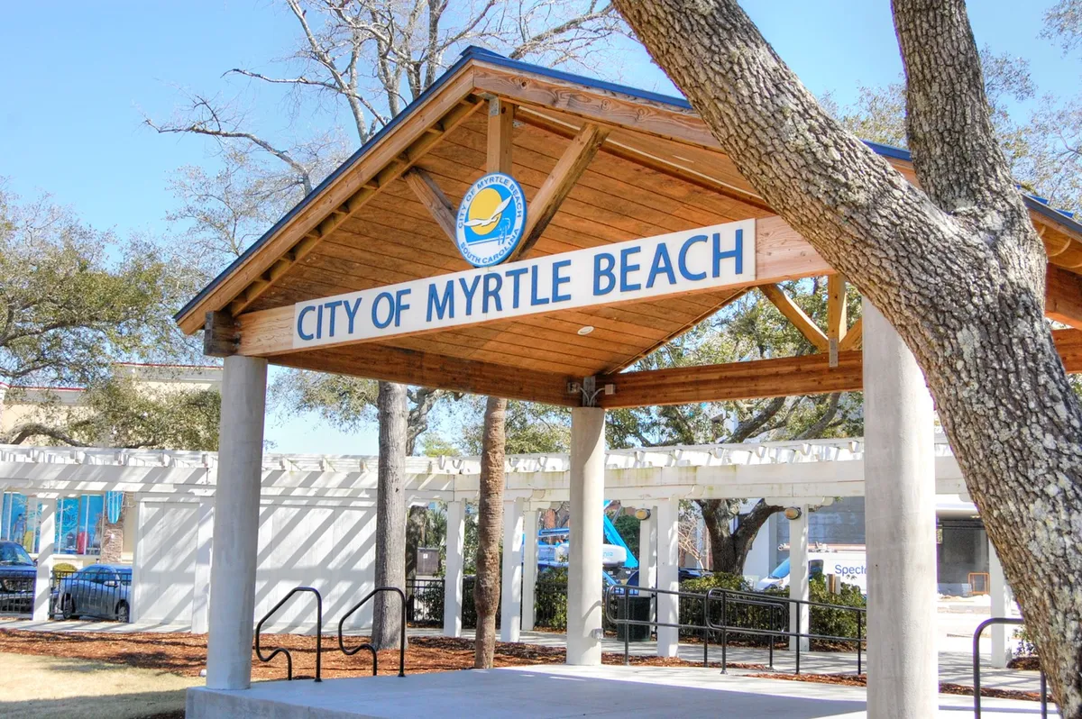A welcoming city sign and wooden structure set among trees, inviting visitors to explore local community spaces. The setting suggests nearby cultural attractions, local events, and a friendly, inviting atmosphere.