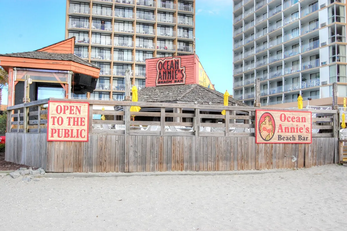 The image showcases the recognizable Ocean Annie’s Beach Bar, complete with signage, a welcoming open structure, and nearby tall buildings. The casual beach bar ambiance and its proximity to the sand set a vibrant, family-friendly scene.