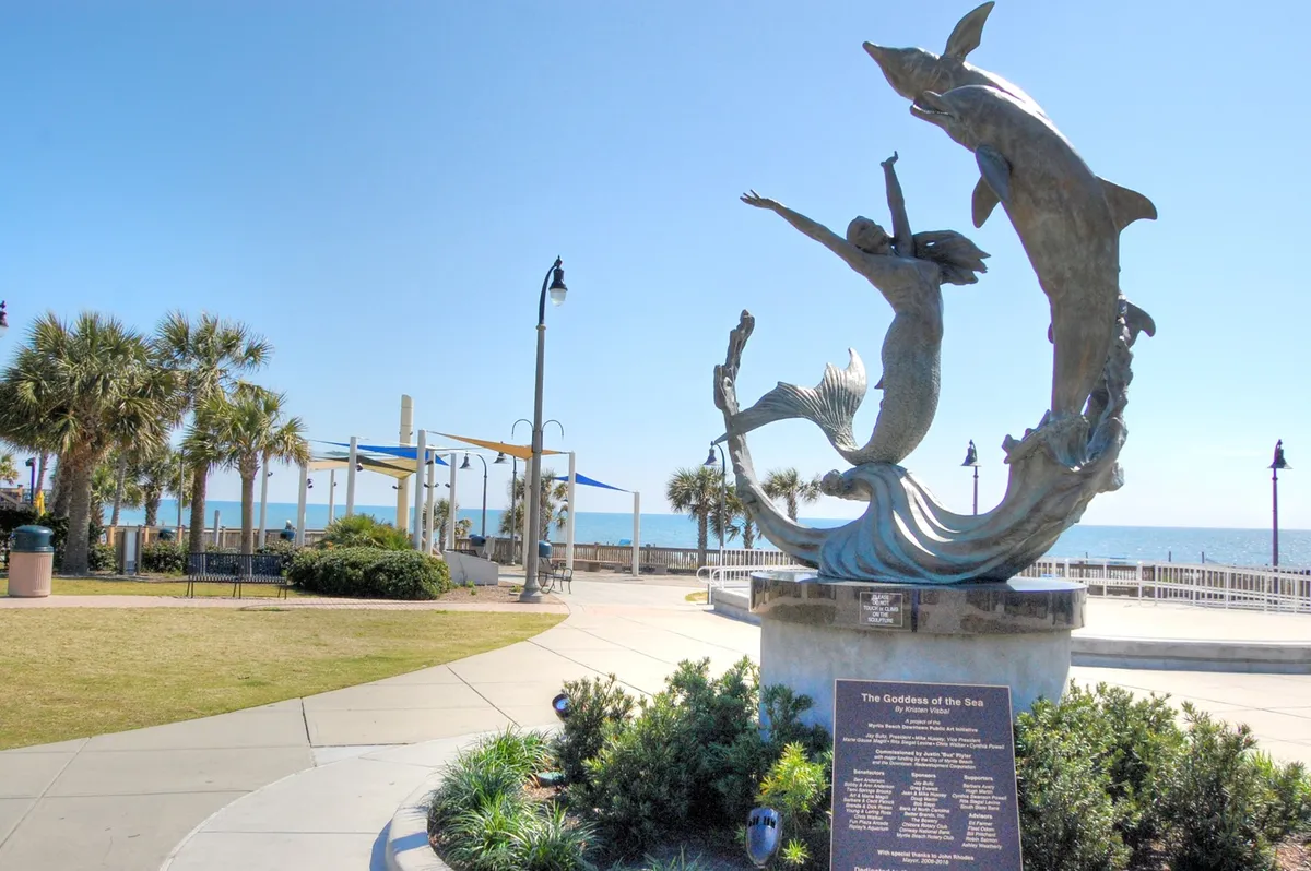 A scenic beachfront park with a dramatic sculpture of the Goddess of the Sea, set against a backdrop of palm trees and ocean views. Couples and visitors leisurely walk the shoreline, pause for photos, and relax on grassy spots, all in a serene, uplifting atmosphere.
