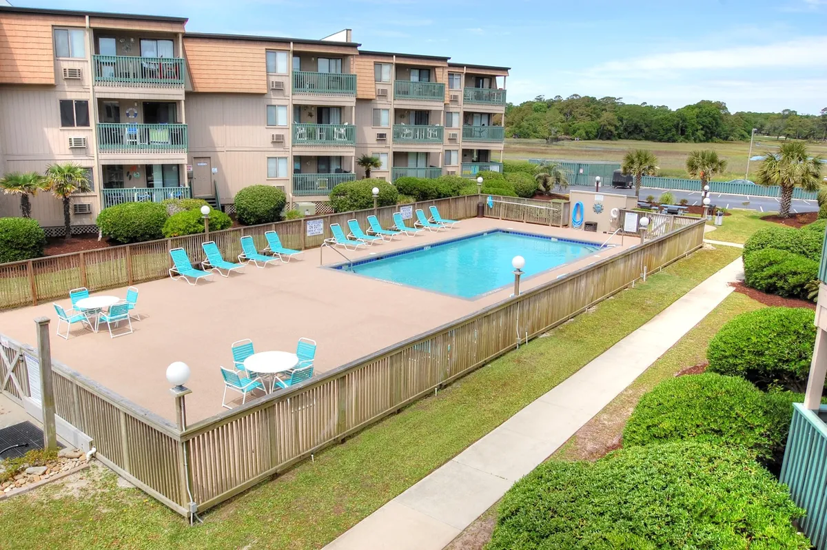 This image displays a sparkling swimming pool surrounded by inviting lounge chairs and lush landscaped greenery. The balconies of the condo building are visible in the background, suggesting the proximity of the accommodations to the resort amenities. The scene exudes a tranquil and inviting mood, with hints of guests relaxing, swimming, and socializing by the pool.
