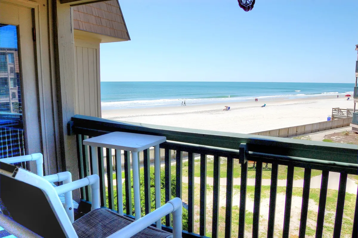 This image offers a stunning view of the Myrtle Beach shoreline as seen from a condo balcony, with comfortable seating positioned to enjoy the tranquil ocean expanse. Sunbathers can be seen on the sandy beach below, and the lively atmosphere of the coastline is visible in the distance.