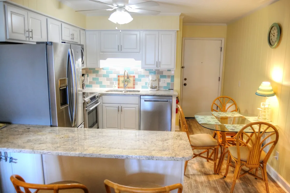 A close-up view of the kitchen area emphasizing the granite countertop, stainless steel appliances, and colorful backsplash, complemented by wooden dining chairs. The scene is lively yet relaxed, suggesting home-cooked meals and quality time around the table.