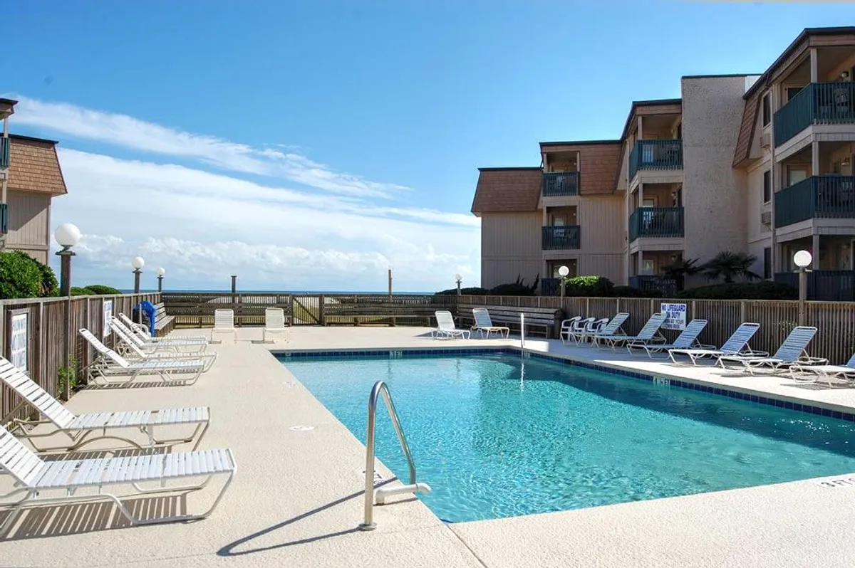 A sunlit swimming pool with lounge chairs frames stunning ocean views beyond. The building’s balconies hint at condo living, while the pool area is alive with leisure, socializing, and relaxation. The atmosphere is leisurely and carefree, perfect for vacationers seeking both excitement and tranquility.
