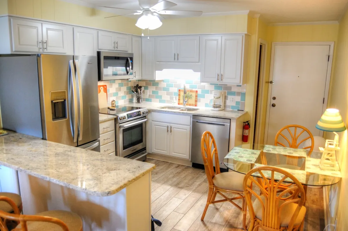 This image features a modern kitchen with a welcoming atmosphere, showcasing a kitchen island with bar stools, a glass dining table surrounded by wicker chairs, and a vibrant backsplash. The setting highlights both the food preparation area and a comfortable space for casual dining and socializing.