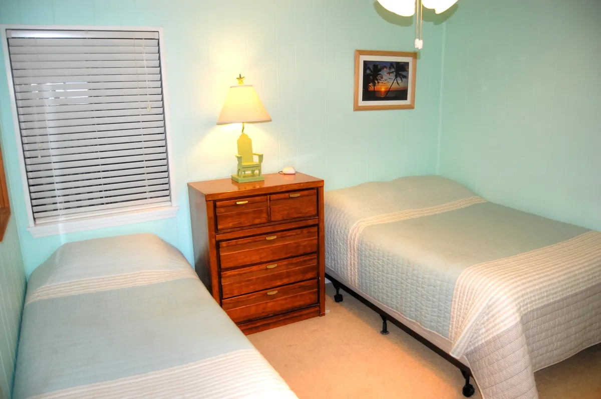 A peaceful second bedroom setup with two beds—one slightly larger than the other—positioned next to a wooden dresser. A table lamp offers cozy lighting, and a framed picture adorns the wall. Gentle morning light filters through the window, creating a welcoming environment for guests of all ages.