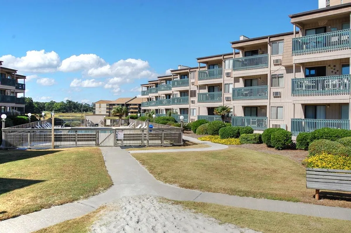 A cheerful, inviting scene of apartment balconies facing landscaped gardens, with a winding walking path and a cozy gated area. People relax on balconies, stroll through the gardens, and gather in small outdoor groups, creating a welcoming, community-focused atmosphere.