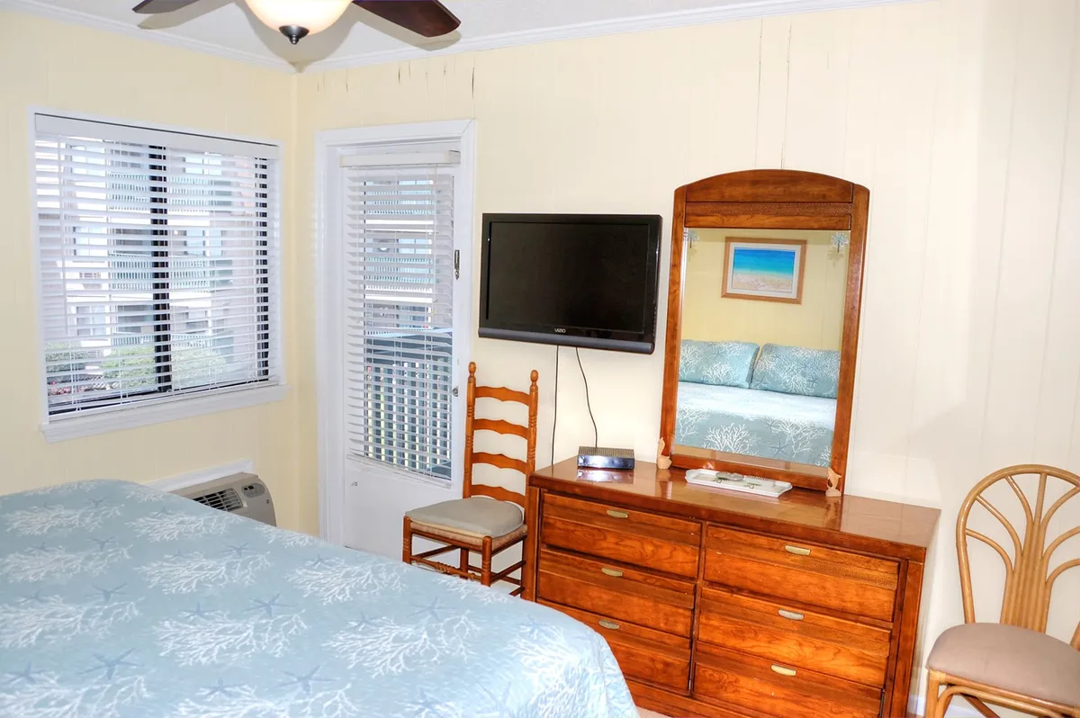A spacious bedroom with a blue quilt on the bed, a mirror with a wooden frame, a dresser with a TV, and a window with blinds allowing in soft light. The mood is calm and serene, with features catering to both relaxation and entertainment.