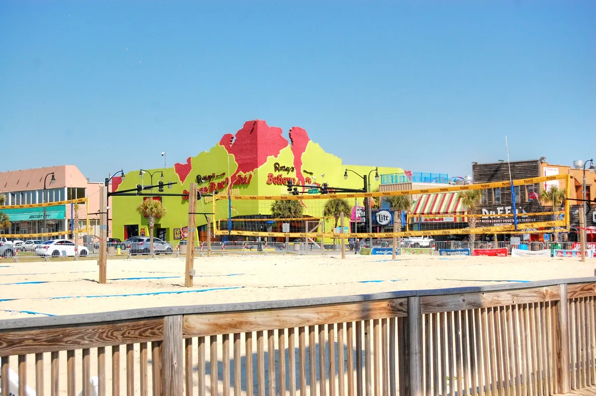 An energetic beachfront scene with a brightly colored building, outdoor seating, and active volleyball courts right on the sand. Groups of friends gather to play, dine, and relax, with the ocean providing a beautiful backdrop.