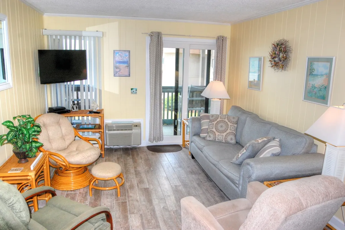 A bright, welcoming living room with a comfortable seating area oriented around a television. Decorative wall art adds a coastal touch. A large sliding glass door opens to a patio, filling the space with natural light and providing easy access to the balcony.