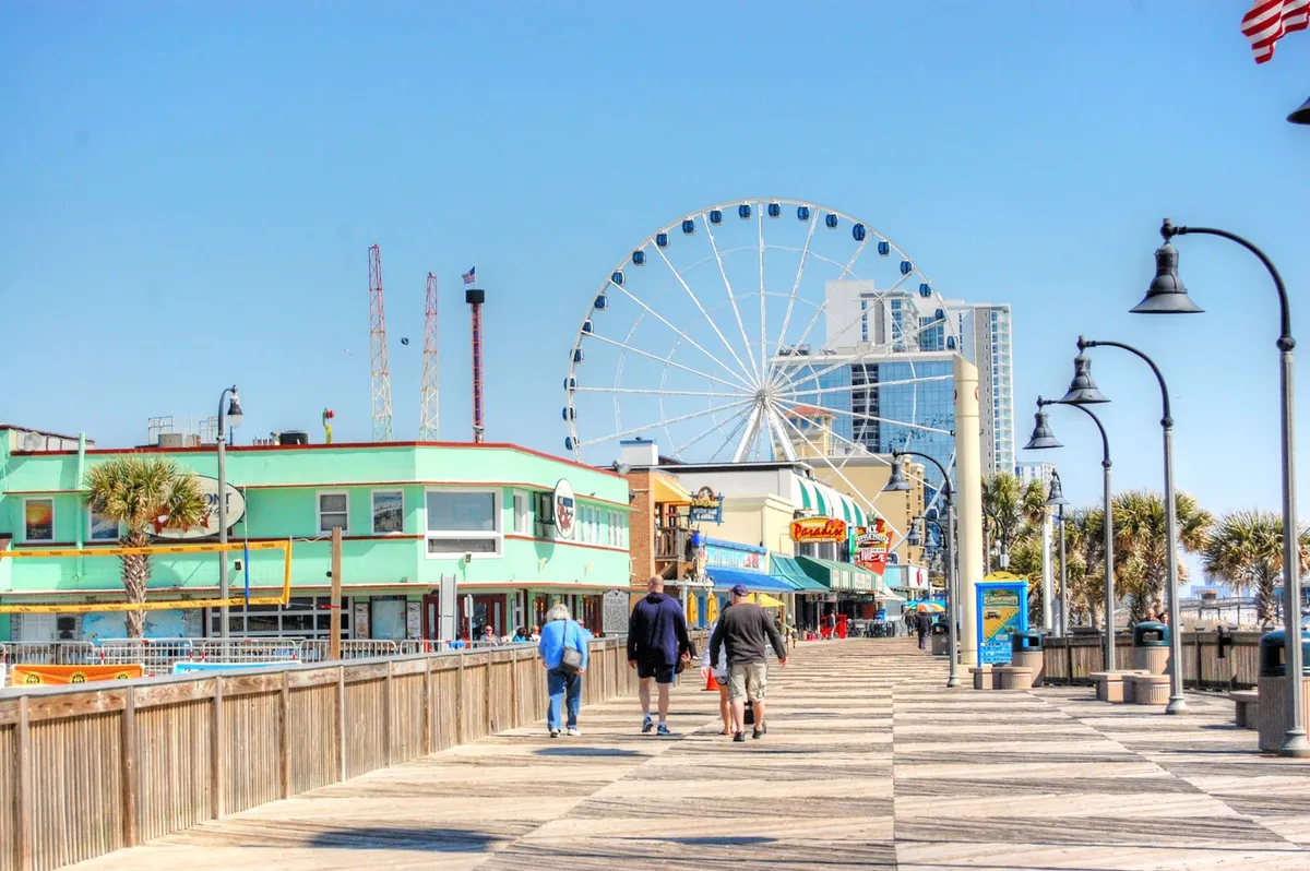 A colorful, energetic view of the Myrtle Beach boardwalk, with its iconic ferris wheel, palm-lined walkways, and a mix of shops and amusements. The scene is filled with people enjoying rides, walking, and shopping, encapsulating the excitement of Myrtle Beach after dark.